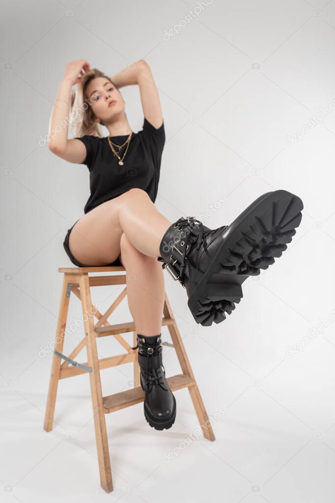 Beautiful fashion girl in black summer clothes with sexy legs in black stylish leather shoes sits on wooden chair. New collection of woman's trendy footwear