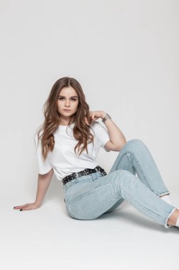 Glamorous beautiful serious girl model in casual clothes with a white t-shirt and blue high waist  jeans sits in the studio