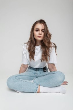 Güzel, kıvırcık saçlı, modaya uygun beyaz tişörtlü, kot pantolonlu ve beyaz çoraplı bir manken kız stüdyoda beyaz arka planda oturuyor.