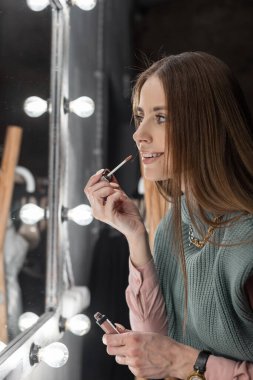 Eski moda elbiselerle gülümseyen mutlu güzel kız aynanın yanında ampullerle dudaklarını boyuyor. Cuma partisi için hazırlanan kadın.