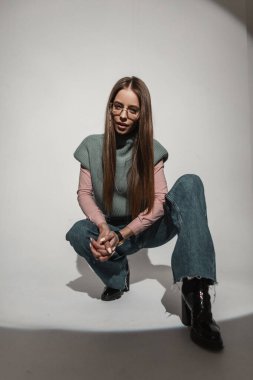 Moda gözlüklü, modaya uygun, eski moda elbiseli, pembe gömlekli, geniş bacaklı, parlak kotlu ve ayakkabılı çekici bir kadın stüdyoda oturuyor. Havalı kız.