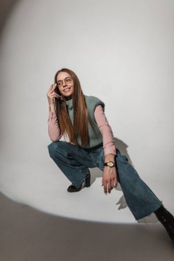Gülümseyen, modaya uygun örgü giysiler içinde eski moda gözlüklü, şık kotlu, ayakkabılı genç ve güzel bir kız model stüdyoda oturur ve poz verir. Klasik tarzda giyinmiş güzel bir kadın.