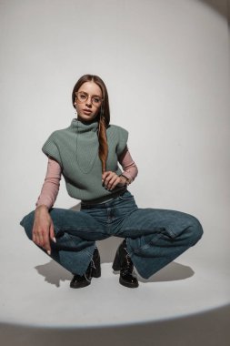 Havalı, şık, altın gözlüklü, kaliteli elbiseli, örgü kazaklı, geniş bacaklı kotlu havalı bir kadın stüdyoda beyaz arka planda oturuyor. Moda kıyafetli güzel bir kız.