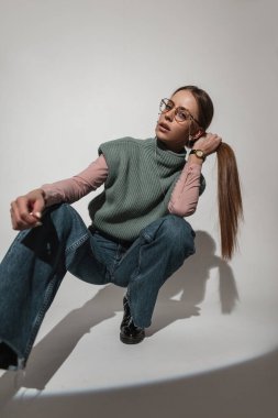 Havalı, şık gözlüklü, şık örülü, geniş kotlu bir yeleği olan genç ve şık bir kız stüdyoda oturuyor ve saçlarını tutuyor. Kapalı alanda güzel bir kadın.