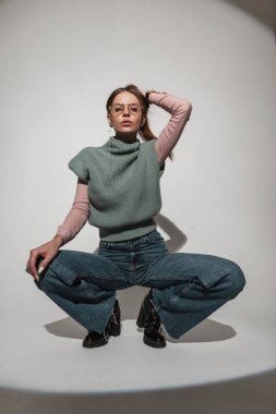 Eski moda gözlüklü, şık giyimli, örgü yelekli, eski moda kotlu ayakkabılı güzel bir kadın stüdyoda oturuyor ve saçlarını tutuyor. Havalı kadın modeli modaya uygun görünüyor.