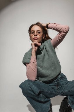 Havalı, genç ve güzel bir manken. Moda kıyafetleri içinde altın sarısı gözlüklü, pembe gömlekli, parlak kotlu beyaz arka planda stüdyoda poz veriyor. Çekici yüzlü modaya uygun bir kadın.