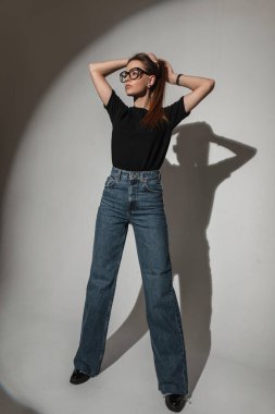 Stylish beautiful glamorous young woman with vintage glasses in a black t-shirt with blue fashion jeans in the studio