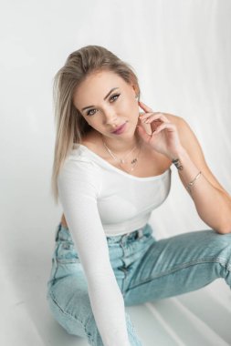 Zarif, genç, sarışın, şirin yüzlü, şık giyimli, kot pantolonlu, beyaz tişörtlü bir kadın stüdyoda oturuyor.