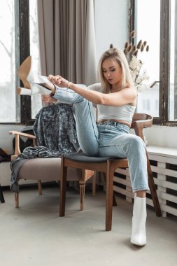 Şık kot elbiseli güzel, genç, sarışın kadın beyaz deri ayakkabılar giyer ve odada bir sandalyeye oturur.