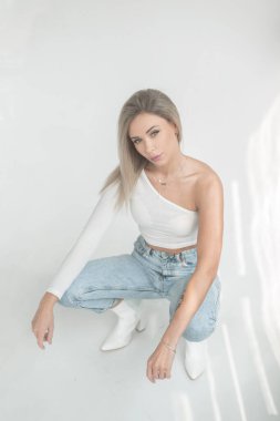 Şık, alımlı, beyaz tişörtlü, kot pantolonlu ve ayakkabılı sarışın kız beyaz bir arka planda oturuyor.