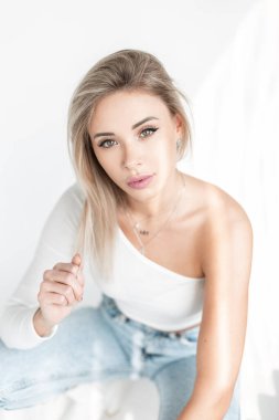 Cute blonde woman in fashion white clothes with t-shirt and blue jeans sits in studio at sunlight