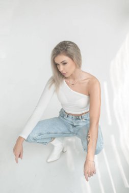 Şık, beyaz tişörtlü, mavi kot ve deri ayakkabılı güzel sarışın kadın gün ışığında stüdyoda beyaz bir arka planda oturuyor.