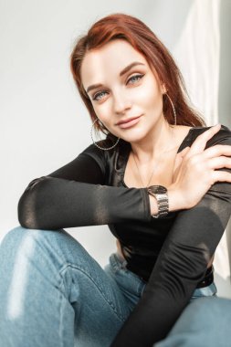 Stylish lovely girl model with beautiful blue eyes in black t-shirt and blue jeans sits indoors at sunlight
