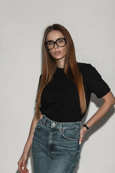 年轻时尚女模特 戴着时髦的眼镜 穿着时髦的黑色T恤和蓝色牛仔裤 站在白墙旁边 穿着时髦衣服的漂亮姑娘 — 图库照片