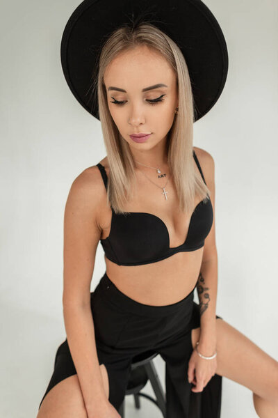 Sexy beautiful young woman in a fashionable black hat with a black bra and pants sits on a chair in the studio on a white background