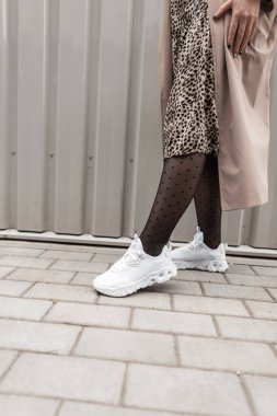 Beyaz spor ayakkabılı, leopar desenli ve ceketli bir kadın metal duvarın yanında sokakta yürüyor.
