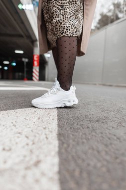 Şık, beyaz spor ayakkabılı, taytlı, leopar giysili, ceketli bir kadın şehirde asfalt üzerinde duruyor.