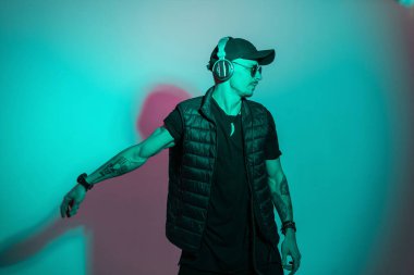 Siyah şapkalı ve güneş gözlüklü dansçı hipster erkek model müzik dinliyor ve stüdyoda yaratıcı pembe cyan renkli ışıkla dans ediyor. Kulaklıklı adam.