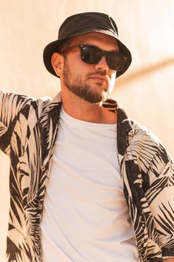 Hipster man with classic sunglasses and a bucket hat in fashionable clothes with a shirt walks on the beach at sunset