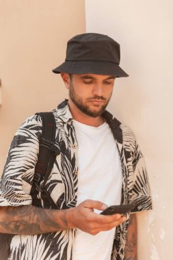 Havai kıyafetleri içinde, bir yaz günü duvarın yanında duran sırt çantası ve telefonu olan şık, yakışıklı bir hippi.