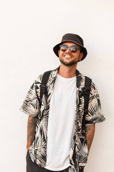 Handsome hipster man with smile in summer fashion clothes with sunglasses, hat and shirt with backpack travel in city