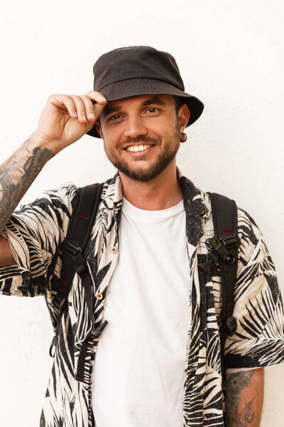 Happy young man model with smile in fashion clothes with hat and shirt stands near white wall
