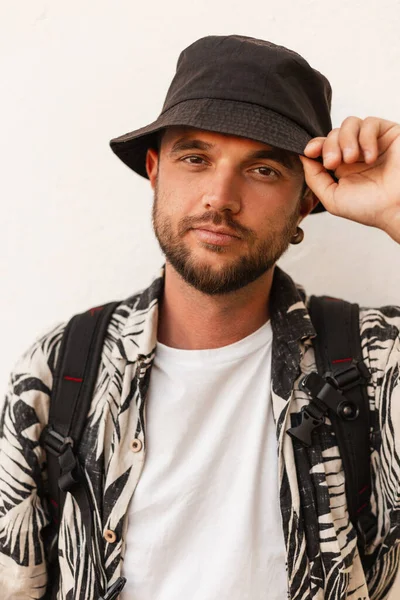 Sırt çantalı şık giyimli yakışıklı hippi şehirde beyaz duvarın yanındaki kova şapkasını düzeltiyor.