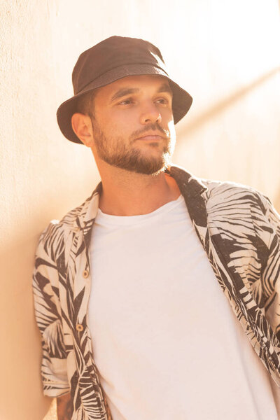 Stylish handsome hipster man with a black panama bucket hat in a fashion beach shirt with palm trees on the beach at sunset