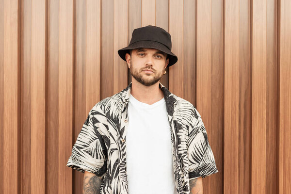 Fashionable handsome hipster man in stylish clothes with Hawaiian shirt, T-shirt and hat standing near the wall