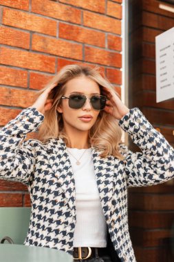 Havalı ceketli şık güneş gözlüklü genç, şık sarışın kadın yaz kafesinde kırmızı tuğlalı duvarın yanında oturur ve dinlenir.