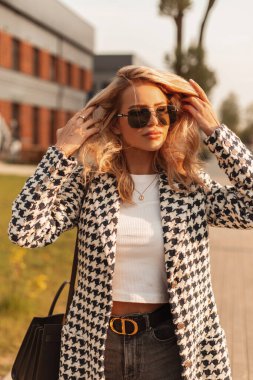 Şık bir hippi şık gözlüklü şık bir sarışın kız şık bir ceket ve el çantalı bir tişört ile gün batımında şehirde yürüyor.