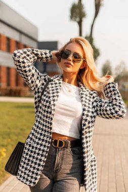 Şık, gösterişli dış giyimli, el çantası ve güneş gözlüklü çekici sarışın kadın gün batımında sokakta yürüyor.