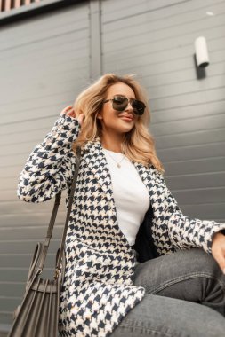 Happy glamorous young girl in fashionable elegant clothes with a jacket, denim, a handbag and sunglasses sits in the city near the modern wall