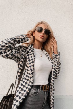 Gözlüklü, şık bir paltosu ve beyaz bir tişörtü olan güzel bir kız sokakta bir duvarın yanında duruyor.