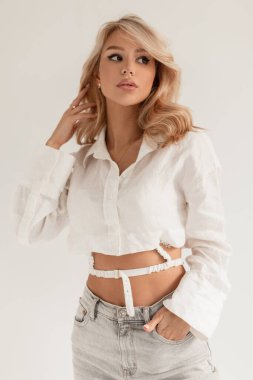 Stüdyoda, beyaz tişörtlü kıvırcık saçlı ve beyaz arka planda yüksek bel kotlu güzel sarışın bir kadının stüdyo portresi.