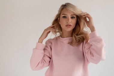 Stylish beautiful blonde model girl in a fashionable pink pullover on a gray background in the studio looks at the camera