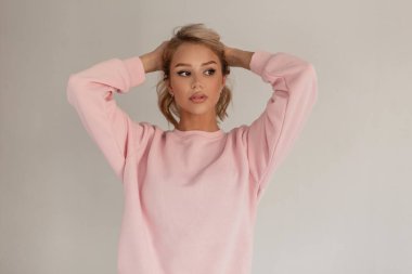 Modaya uygun genç kız model sarışın şık pembe kazaklı, gri arka planda saçlarını düzleştiriyor.