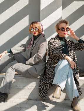 Moda retro giysileri içinde güneş gözlüklü iki şık çekici kız sokağın kenarında güneş ışığıyla beton bir duvarın yanında oturuyorlar. Kadın şehir tarzı kıyafet.