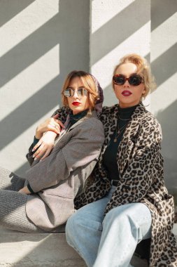 Eski moda güneş gözlüklü, şık retro kıyafetli iki moda güzel kız güneş ışığında sokağın ortasında bir duvarın yanında oturuyor. Şehir modası kıyafetleri.