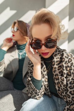 Modaya uygun güneş gözlüklü, leopar desenli ceketli, kot pantolonlu ve kazaklı şık, güzel bir kadın sokakta kız arkadaşıyla oturuyor. Güzel kız kameraya bakar.