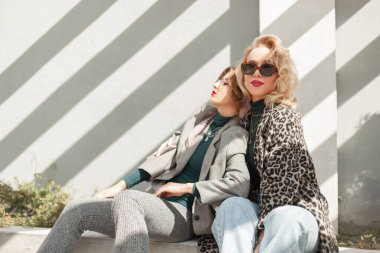 Güneş gözlüklü, leopar desenli ceketli, kot pantolonlu ve takım elbiseli iki modacı kız gün ışığında sokakta oturuyor.