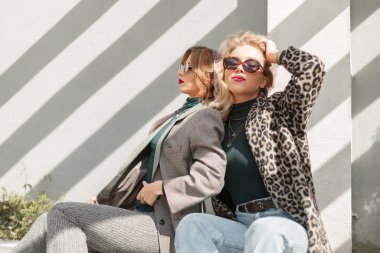 Moda gözlüklü, modaya uygun iki güzel ve şık kız model güneş ışığında sokakta oturur ve dinlenir. Kadın şehir tarzı kıyafetler.