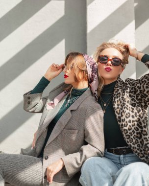 Moda leoparı ceketli, takım elbiseli, kotlu ve kazaklı havalı güneş gözlüklü iki hippi kız gün ışığında sokakta oturuyor.