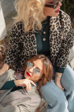 İki şık, komik, güneş gözlüklü, şık elbiseli, leopar desenli, kot pantolonlu, kazaklı ve spor ceketli genç kız. Şehirdeki en iyi moda kız arkadaşları, kadınsı şehir tarzı.