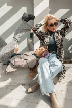 Moda kıyafetleri içinde, leopar desenli, takım elbiseli, kot pantolonlu ve ayakkabılı iki göz alıcı şık kız arkadaş şehirde oturuyor ve poz veriyorlar.