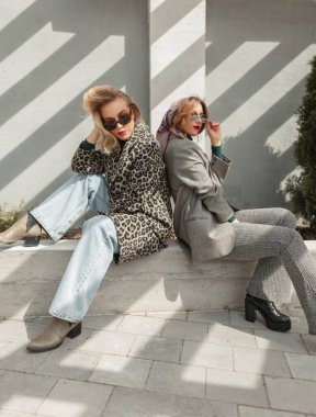 Güneş gözlüğü, leopar desenli ceket, kot pantolon, şık takım elbiseli ve ayakkabılı iki moda kızı şehirde oturuyor.
