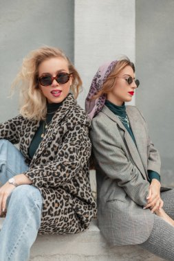 Moda kıyafetleri içinde güneş gözlüklü iki güzel moda hippi kız sokakta oturuyor.