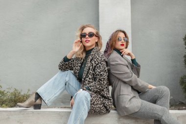 Güneş gözlüklü, modaya uygun leopar desenli ceketli, kot pantolonlu, gri takım elbiseli ve ayakkabılı genç çiftler gri bir duvarın yanında sokakta oturuyorlar.