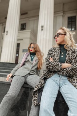 Moda kıyafetleri içinde, moda leopar ceketli, mavi kotlu, gri takım elbiseli ve güneş gözlüklü iki güzel kadın şehirde eski bir binanın yanında oturuyor.