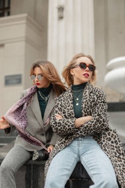 Şık giyinmiş iki güzel genç kız şehirde bir binanın yanında oturuyor.
